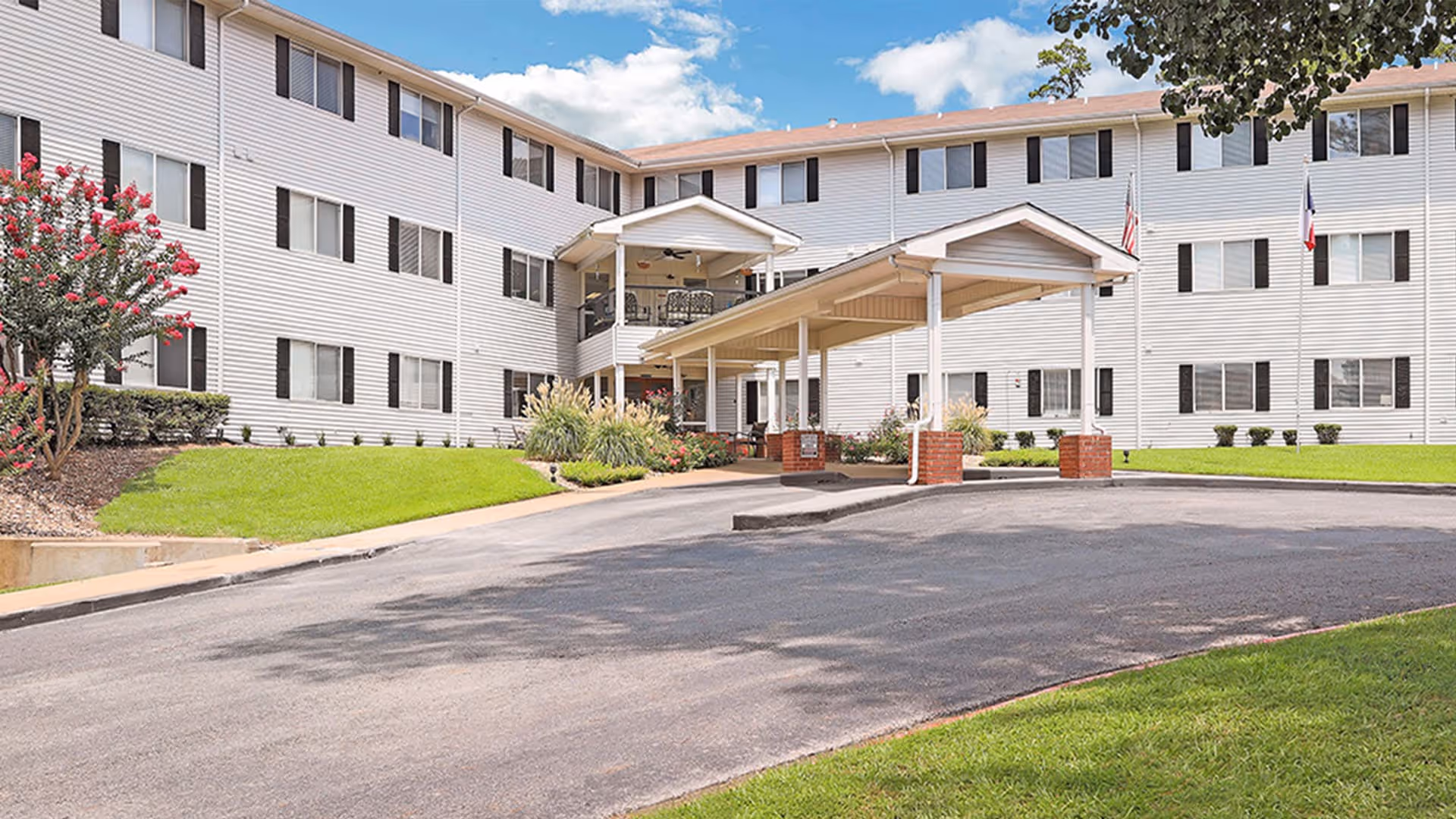 Exterior view of a three-story senior living facility building with white siding and black window shutters. The entrance features a covered driveway with brick pillars and a small landscaped area with green grass, bushes, and flowering plants. Two flags are visible near the entrance under a partly cloudy sky.