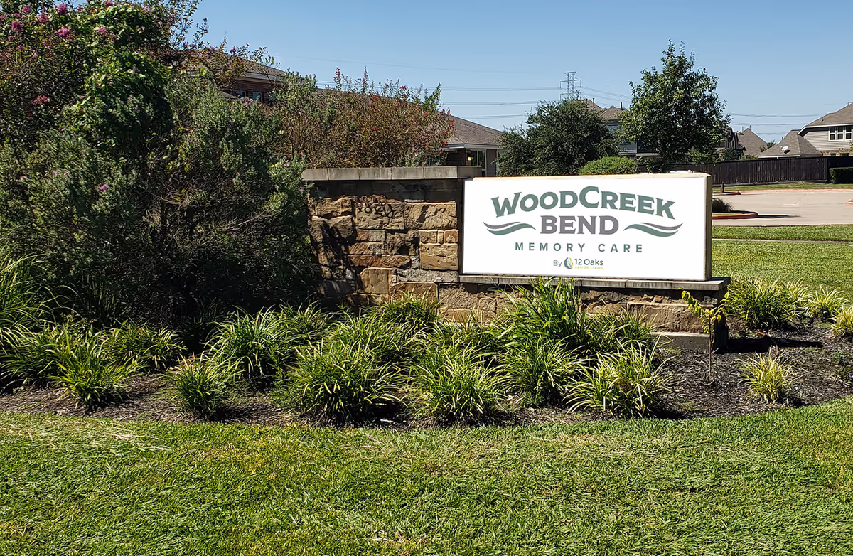 Outdoor stone sign for WoodCreek Bend Memory Care surrounded by green grass, plants, and bushes with residential houses and a clear blue sky in the background.