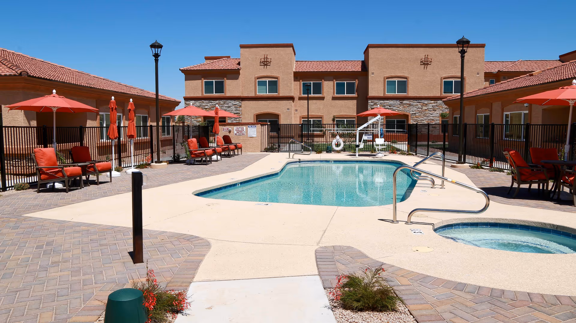 Outdoor swimming pool area at a senior living facility with red cushioned lounge chairs and umbrellas around the pool. The building in the background has a beige exterior with red tile roofing under a clear blue sky.