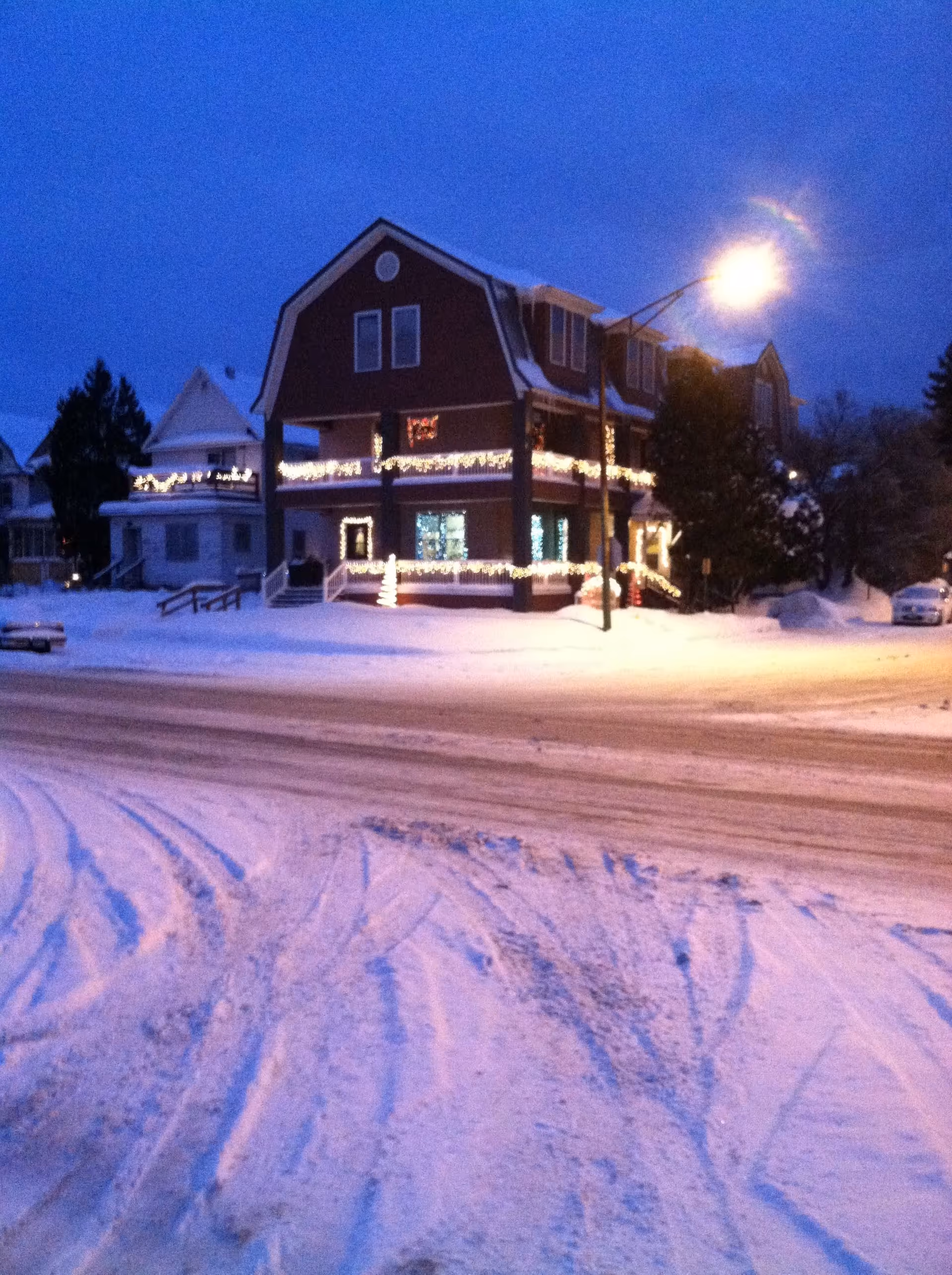 A large two-story house decorated with white Christmas lights on the railings and windows, surrounded by snow-covered ground and streets during early evening or dusk. A streetlamp is lit near the house, and neighboring houses are visible in the background.