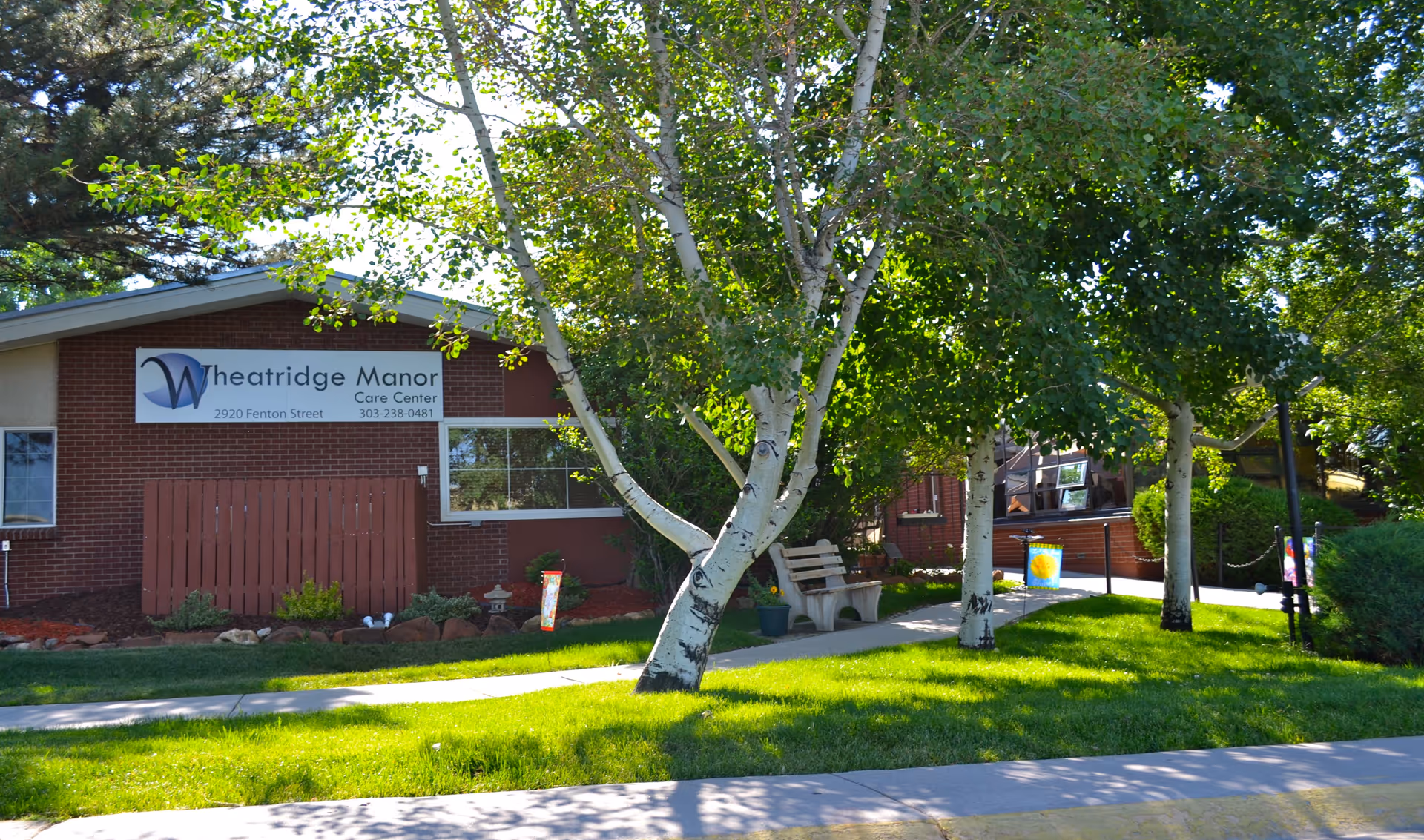 Exterior view of Wheatridge Manor Care Center with sign and trees.