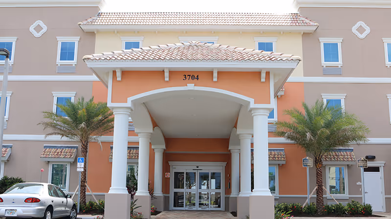 Front entrance of a peach-and-beige multi-story senior living building with a covered porte-cochère, white columns, palm trees, and the number 3704 above the doors.