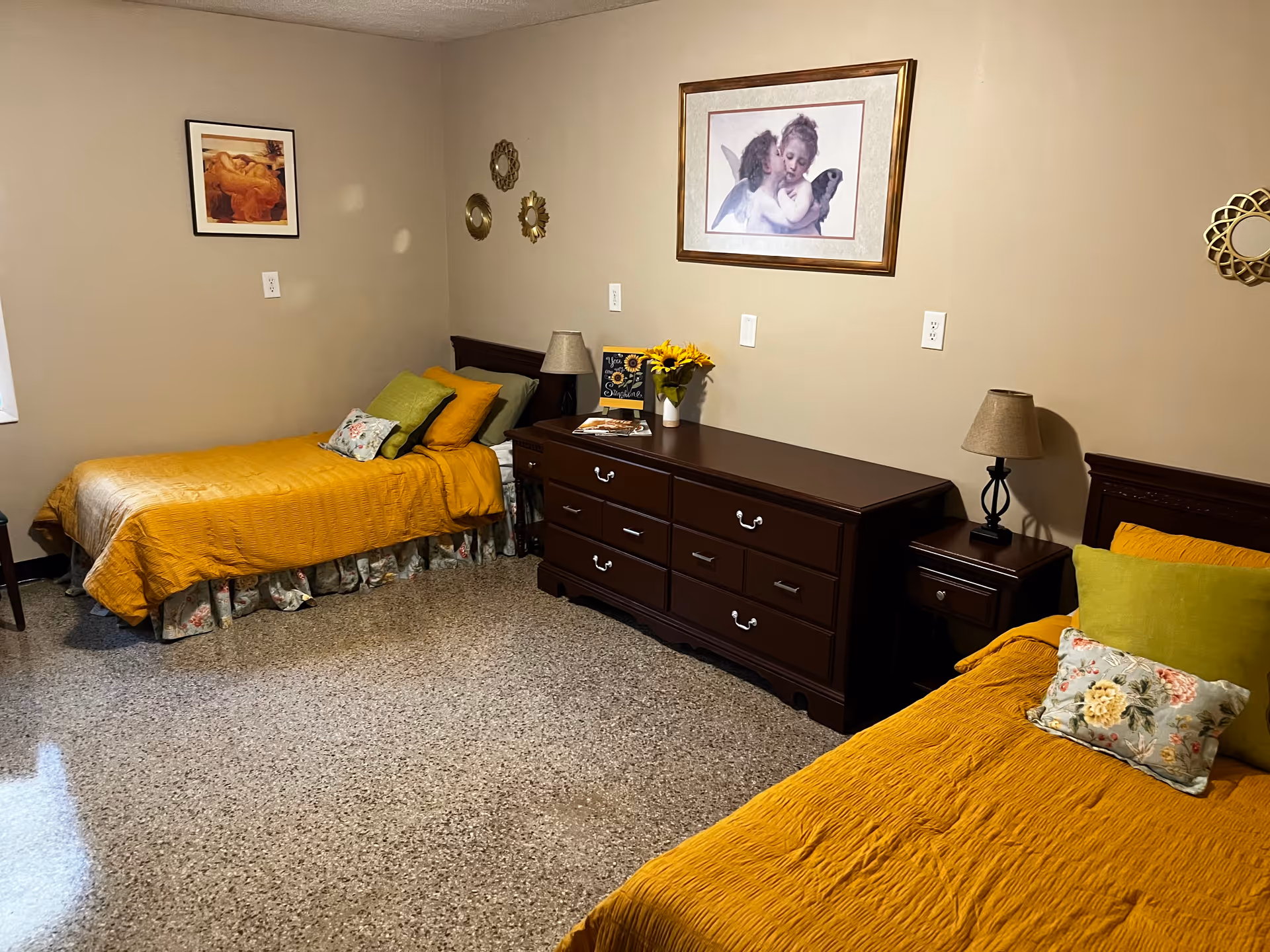 A cozy bedroom with two single beds covered in mustard yellow bedding and floral pillows. Between the beds is a dark wooden dresser with a vase of sunflowers, a small lamp, and decorative items. The walls are beige and decorated with framed artwork and small round mirrors.