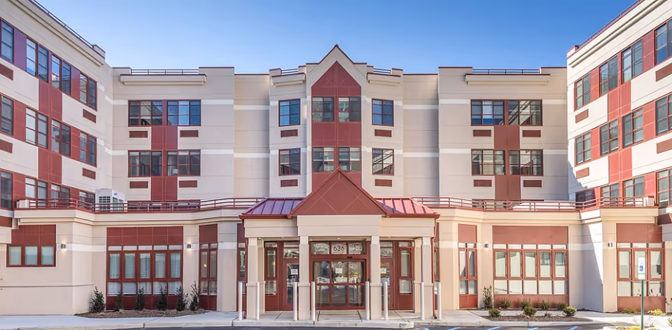 Front entrance and façade of a multi-story senior living facility with red accents and large windows.