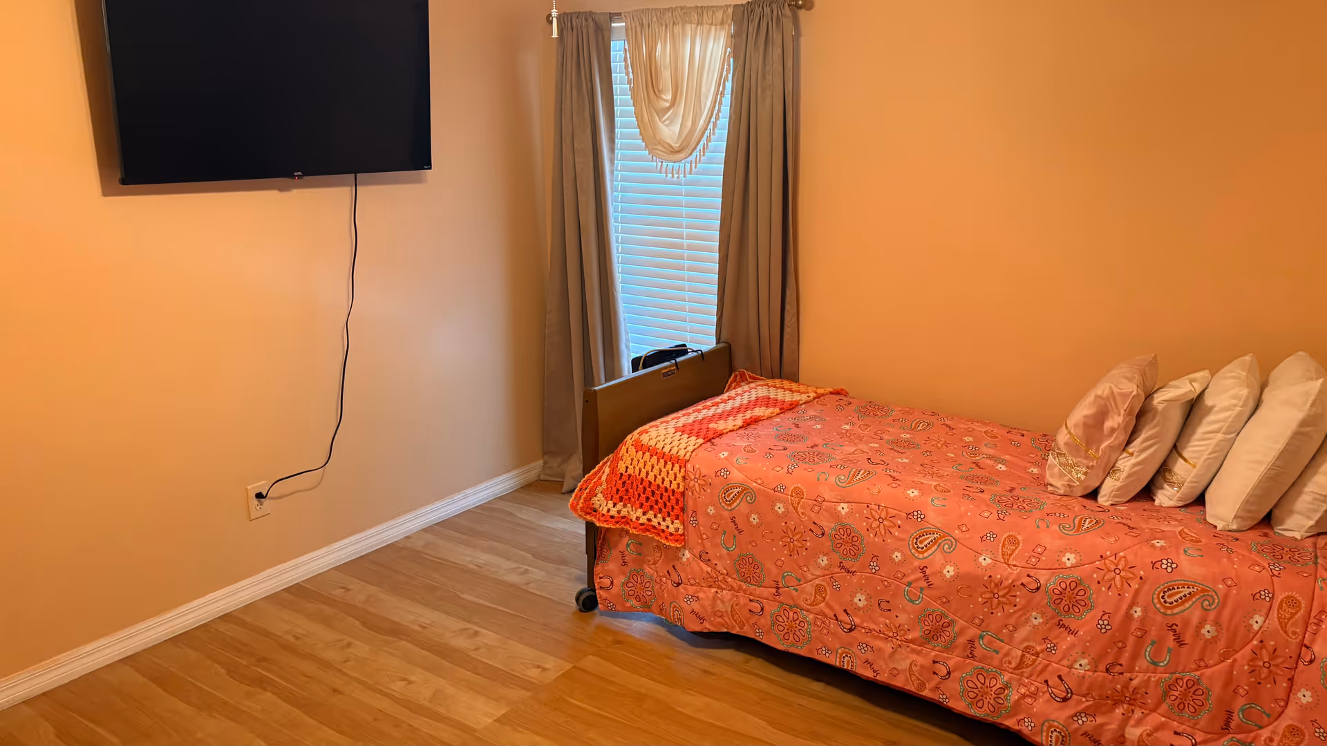 A small bedroom with a pink floral bed, window curtains, and a wall-mounted TV.