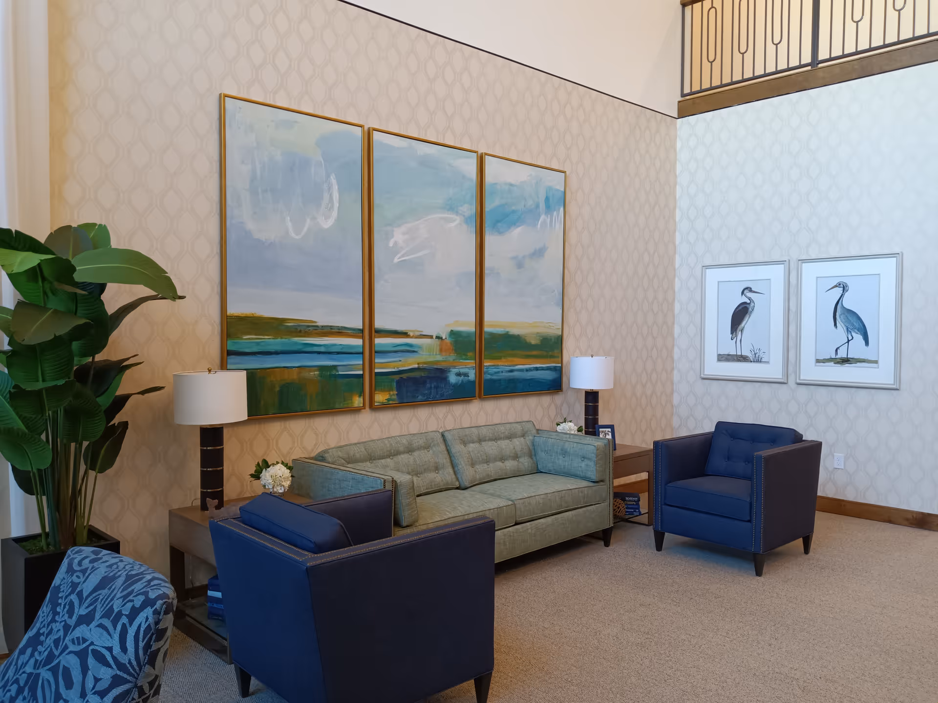 Modern lounge area with green sofa, blue chairs, lamps, plant, and abstract wall art.
