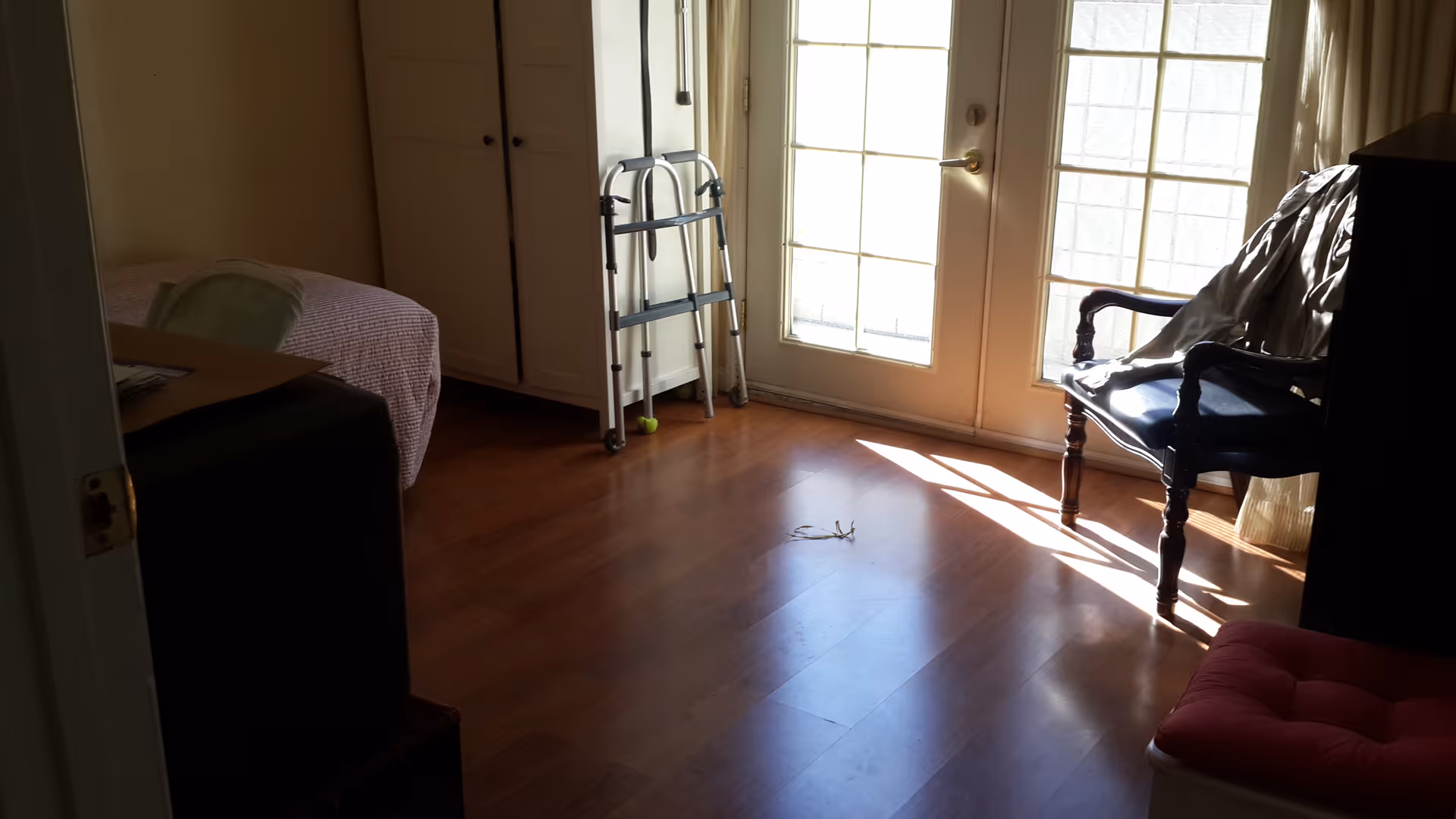 A room with wooden flooring featuring a bed with a pink blanket on the left, a walker near a set of glass double doors letting in sunlight, a dark wooden chair with a light-colored cloth draped over it, and a red cushioned stool in the foreground.