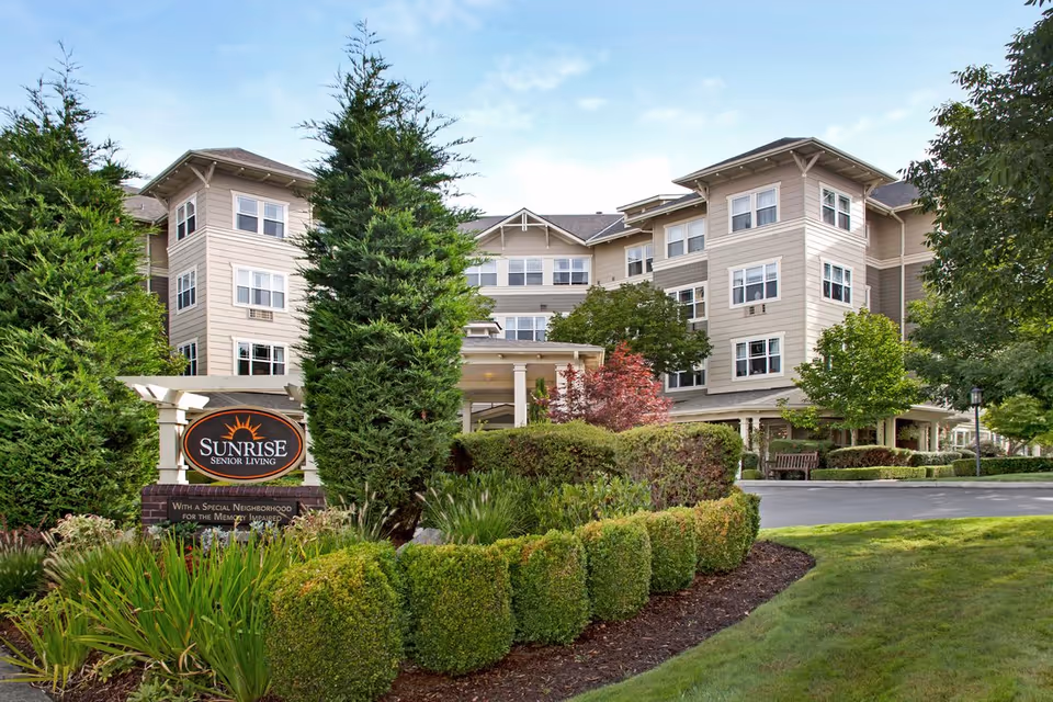 Front exterior of a multi-story senior living building with landscaped entrance and a 'Sunrise Senior Living' sign.