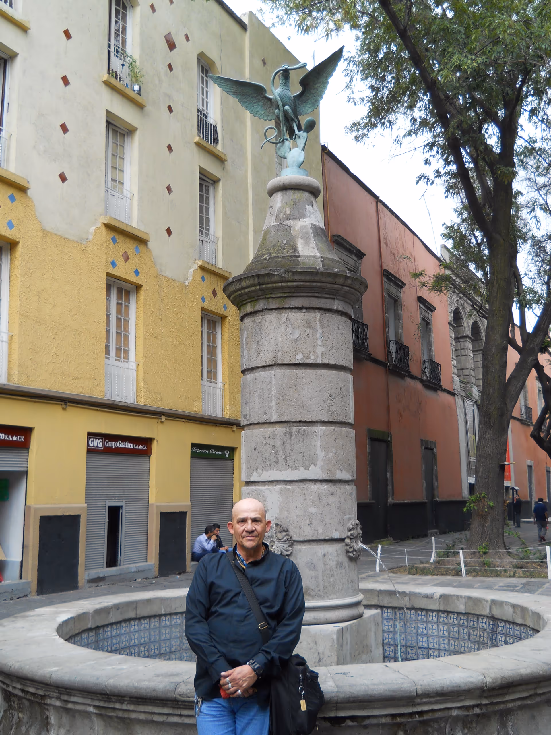 A man stands beside a stone fountain with a winged sculpture, near colorful buildings.