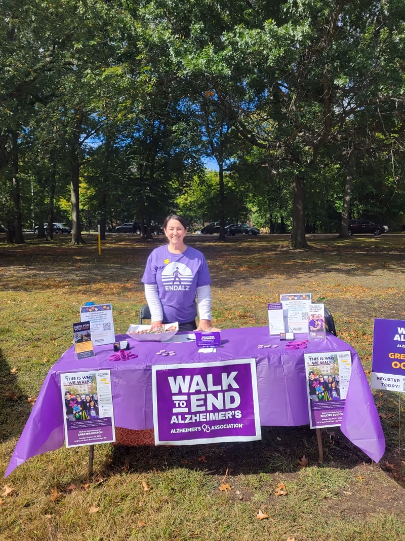 Person in purple shirt stands behind a Walk to End Alzheimer's table outdoors in a park