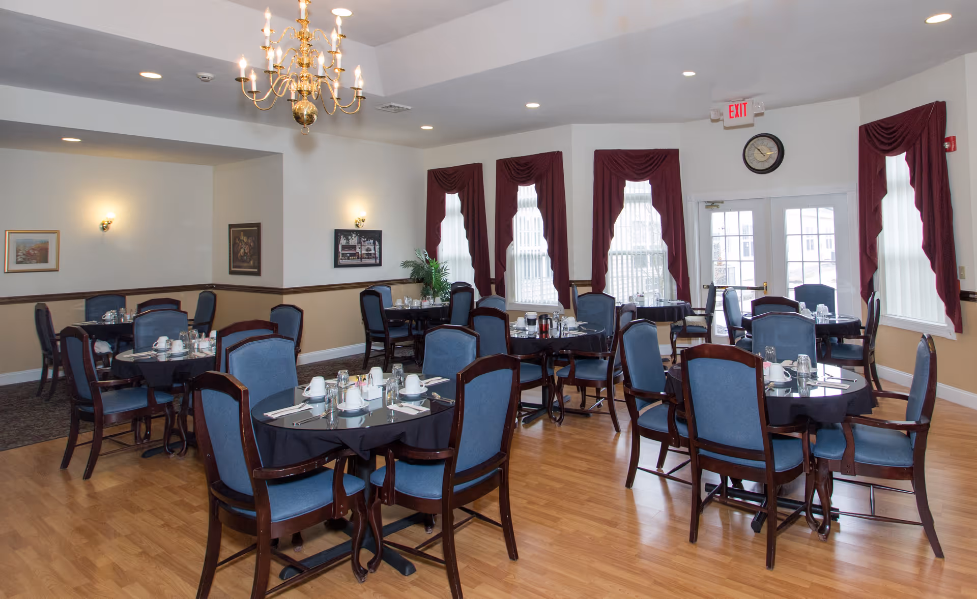 A dining room with several round tables covered with black tablecloths, each set with white cups, plates, and silverware. The room has wooden flooring, blue cushioned chairs with dark wooden frames, burgundy curtains on the windows, a chandelier hanging from the ceiling, and framed artwork on the walls.