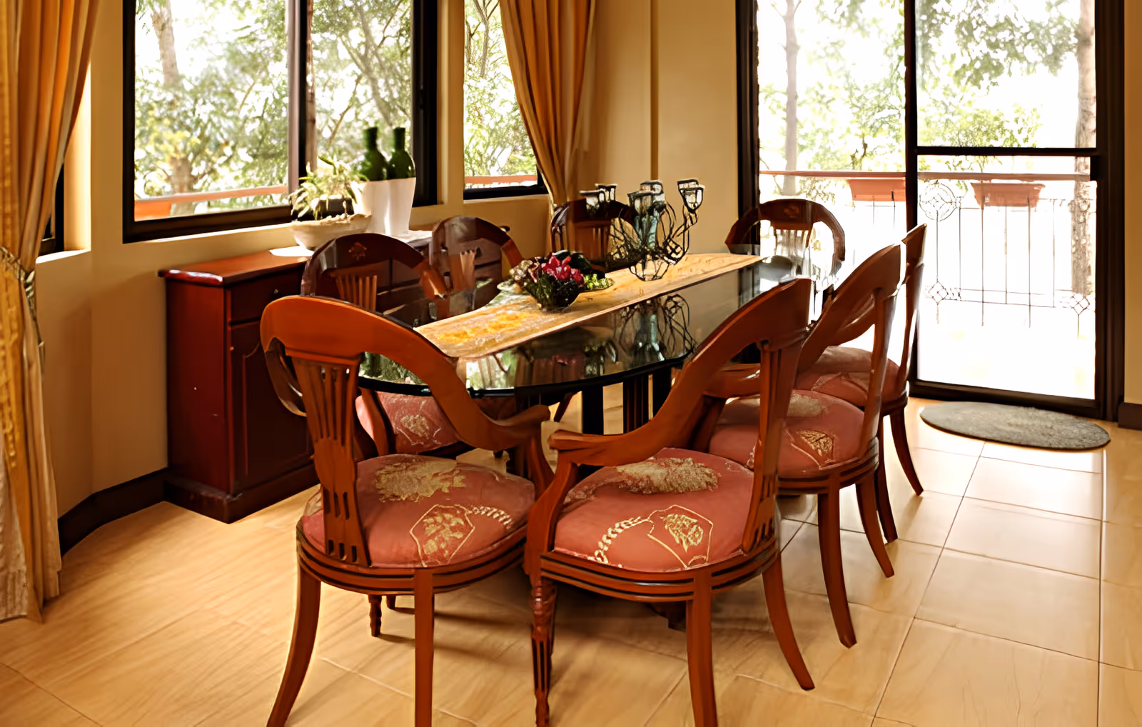 Sunlit dining room with a glass-top table surrounded by wooden upholstered chairs and a sliding door to a balcony.