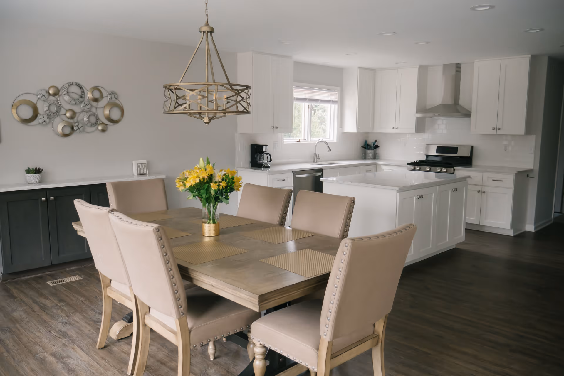 A modern kitchen and dining area featuring a wooden dining table with six beige upholstered chairs, a vase with yellow flowers as a centerpiece, white cabinetry, a kitchen island with a marble countertop, stainless steel appliances including a stove and dishwasher, a coffee maker on the counter, and a decorative wall piece with circular designs.