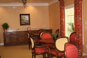 A cozy interior room with a wooden table surrounded by red and cream upholstered chairs. There is a wooden sideboard against the wall with a framed painting above it, a small plant, and decorative items. The room has beige walls, a window with red patterned curtains, and a potted plant near the window.