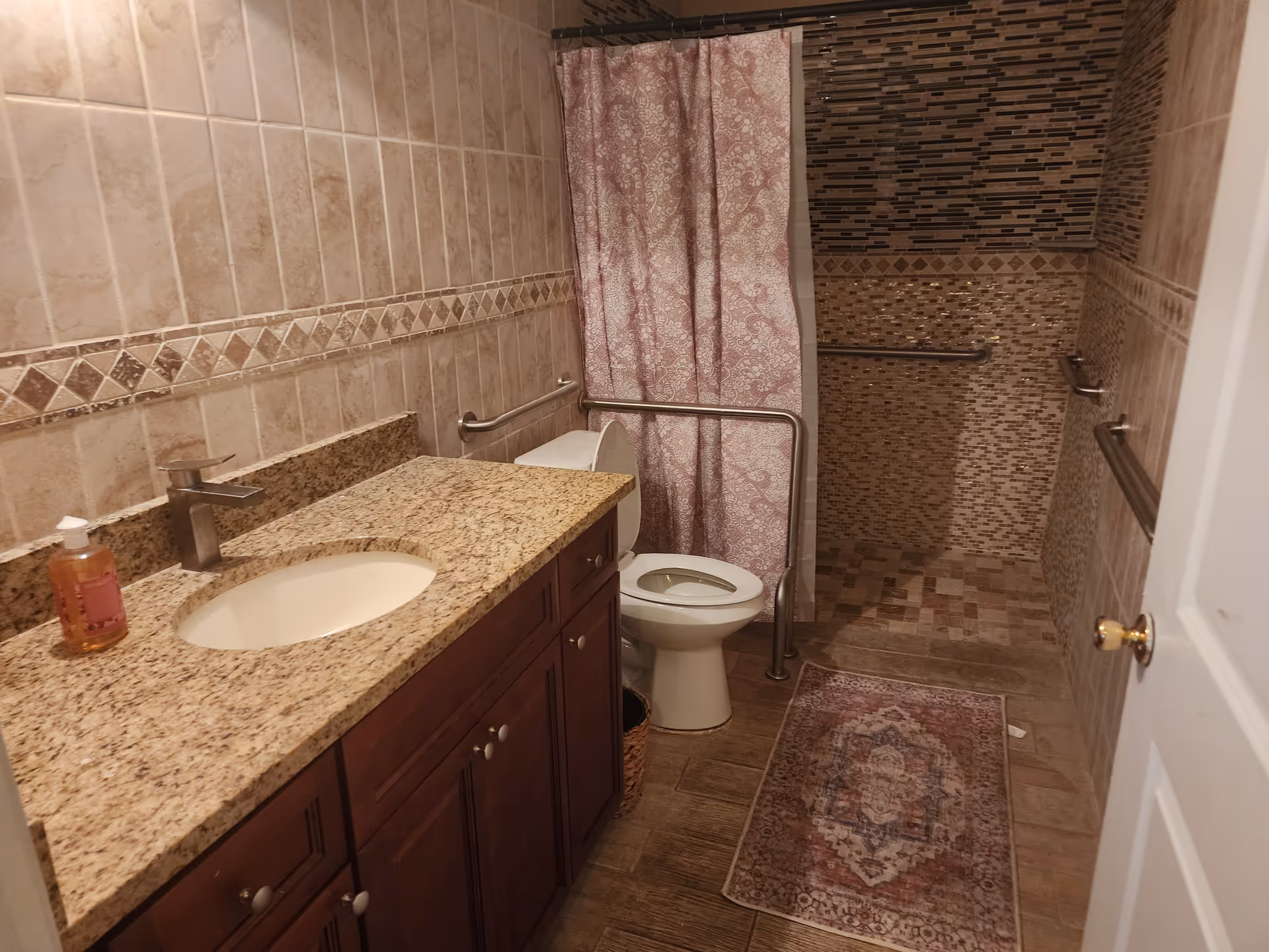 Bathroom with granite vanity, toilet, shower, pink curtain, and grab bars.
