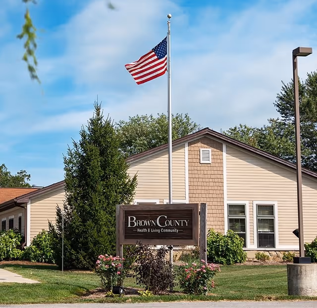 Front exterior of Brown County Health & Living Community showing the building facade, a wooden sign, an American flag, and landscaped shrubs.