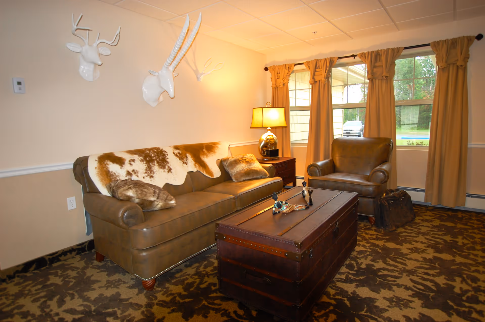 Cozy living room with a leather sofa and armchair, a trunk-style coffee table, decorative white antler mounts on the wall, and a lamp by curtained windows.