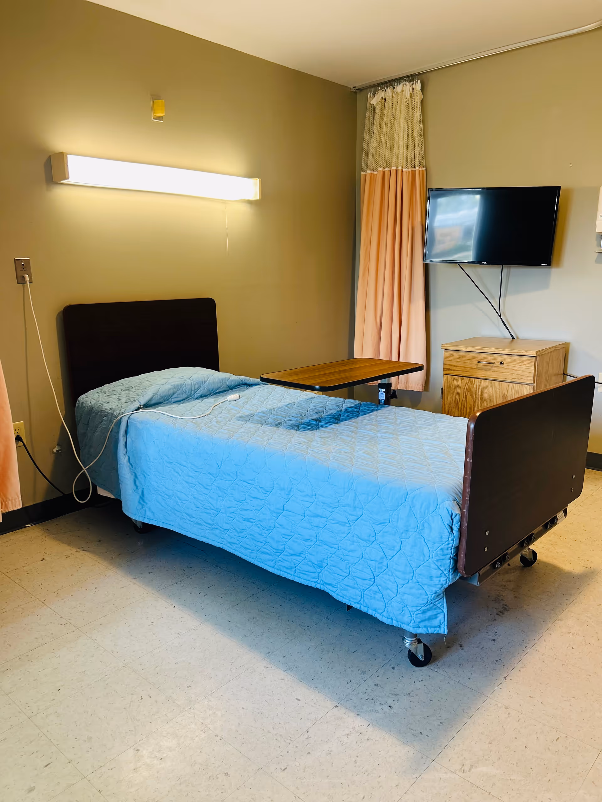 A single hospital-style bed with a blue quilted blanket in a room. The bed has a wooden headboard and footboard and is on wheels. There is a wooden overbed table positioned over the bed. A wall-mounted flat-screen TV is visible on the right side above a wooden cabinet. A peach-colored curtain hangs in the corner, and a long fluorescent light fixture is mounted on the wall above the bed.