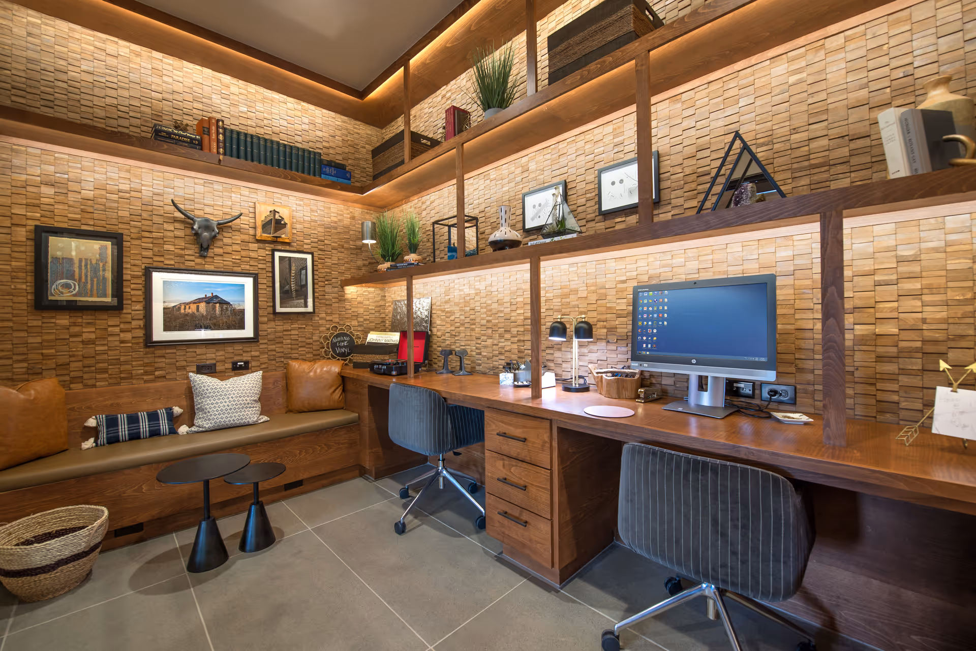 A cozy office or study area with a wooden desk, two gray office chairs, and built-in wooden shelves filled with books, plants, and decorative items. The walls have a textured wooden panel design. There is a cushioned bench with pillows along one wall, two small black side tables, and a computer monitor on the desk.