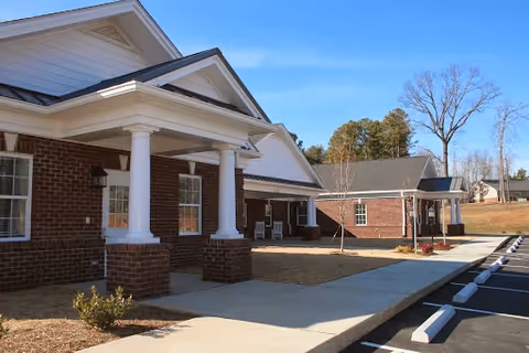 Exterior view of a brick building with white columns and a covered entrance, a sidewalk leading to the entrance, a parking lot with marked spaces, and trees in the background under a clear blue sky.