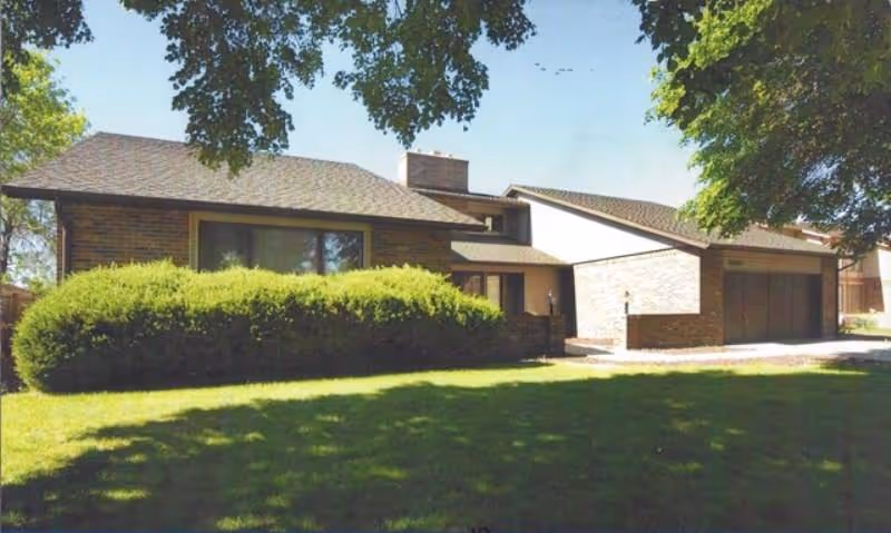 Exterior view of a single-family house with a sloped roof, large windows, and a garage. The house is surrounded by green grass, bushes, and trees under a clear blue sky.