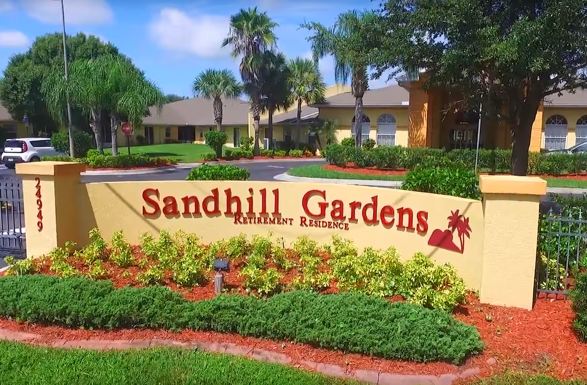 Entrance sign for Sandhill Gardens Retirement Residence with landscaping including green bushes, yellow plants, and palm trees, and a building in the background under a blue sky with some clouds.
