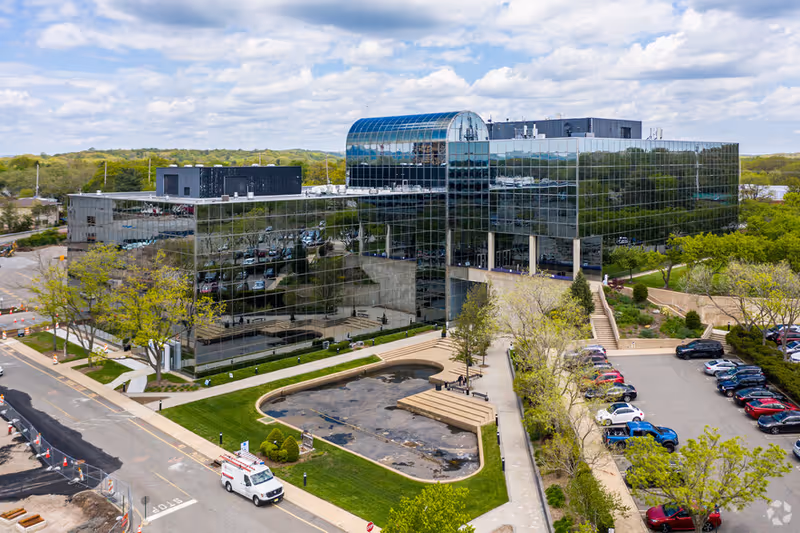 Aerial view of a modern glass office building with surrounding parking and greenery.