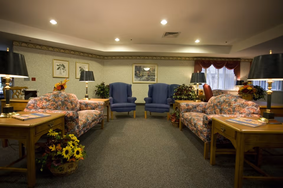 Interior senior living lounge with floral sofas, blue armchairs, lamps, and plants.