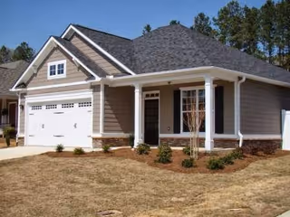 Single-story house with gray siding, white trim, a two-car garage, and a small front porch with white columns. The yard has dry grass and some small shrubs, with trees visible in the background.