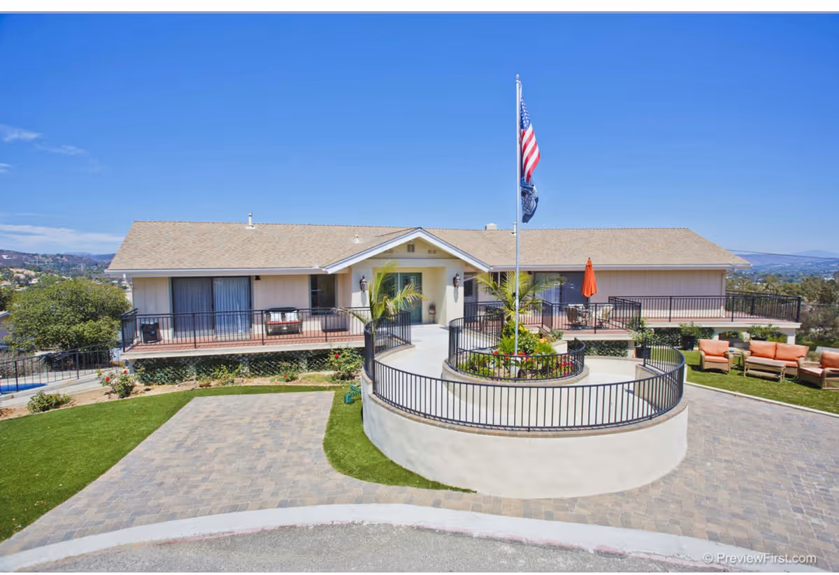 Exterior view of a single-story senior living facility building with a beige roof and light-colored walls. The entrance features a circular raised garden bed with an American flag and another flag on a flagpole. There are outdoor seating areas with chairs and an umbrella on the right side, and the building is surrounded by a paved driveway and green landscaping under a clear blue sky.