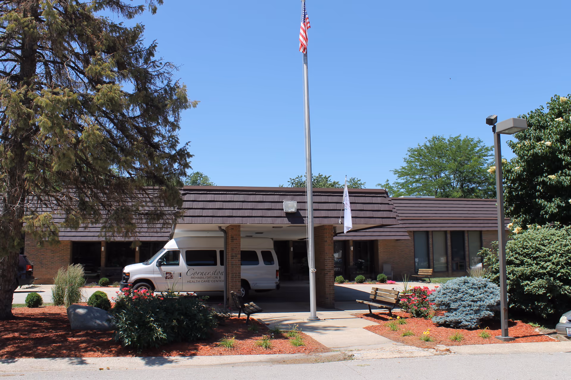 Exterior view of a single-story brick building with a covered entrance. A white van is parked under the entrance canopy. There is a flagpole with an American flag in front of the building, surrounded by landscaped bushes, flowers, and trees. The sky is clear and blue.