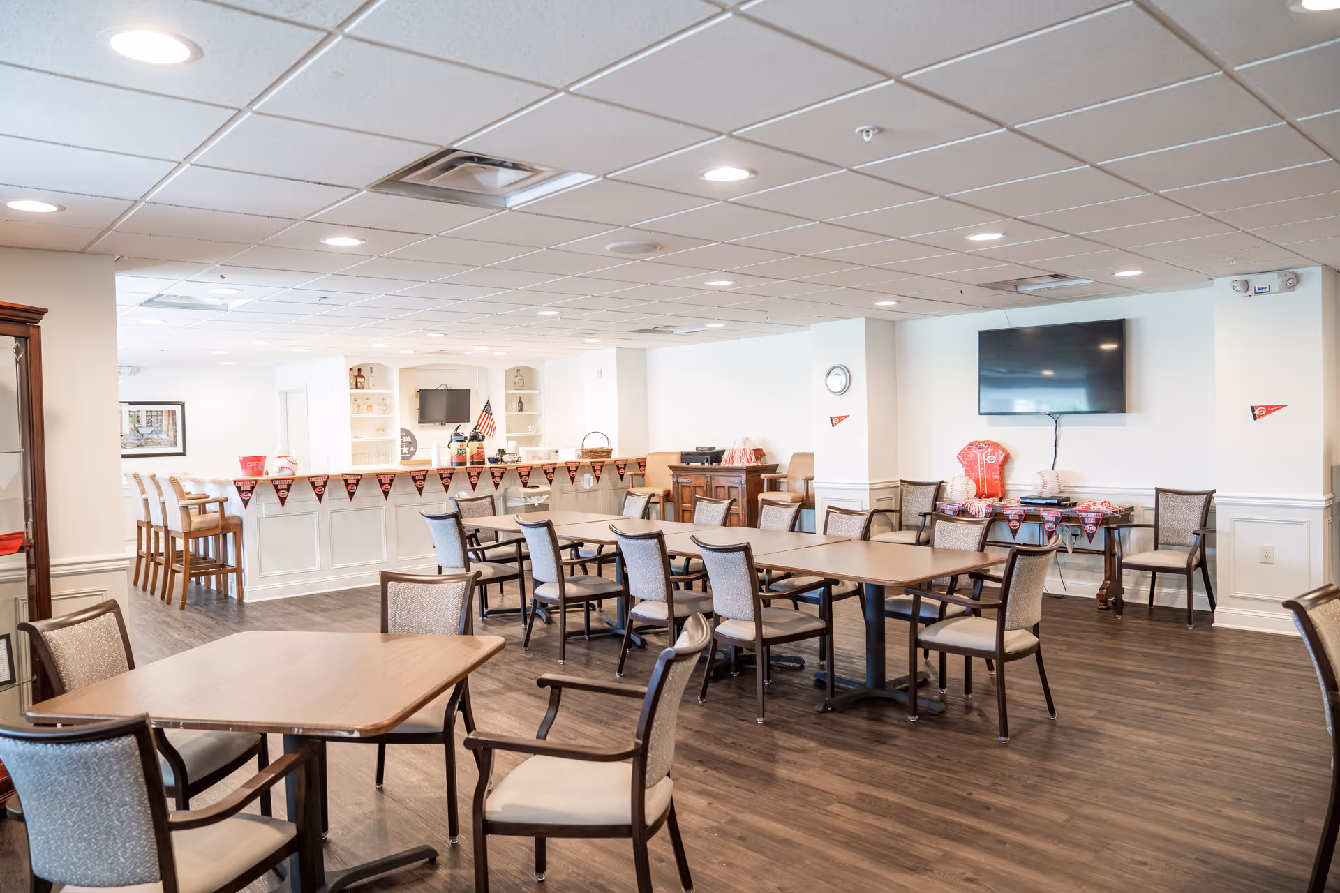 Senior living dining room with multiple tables, chairs, and a serving counter in background.