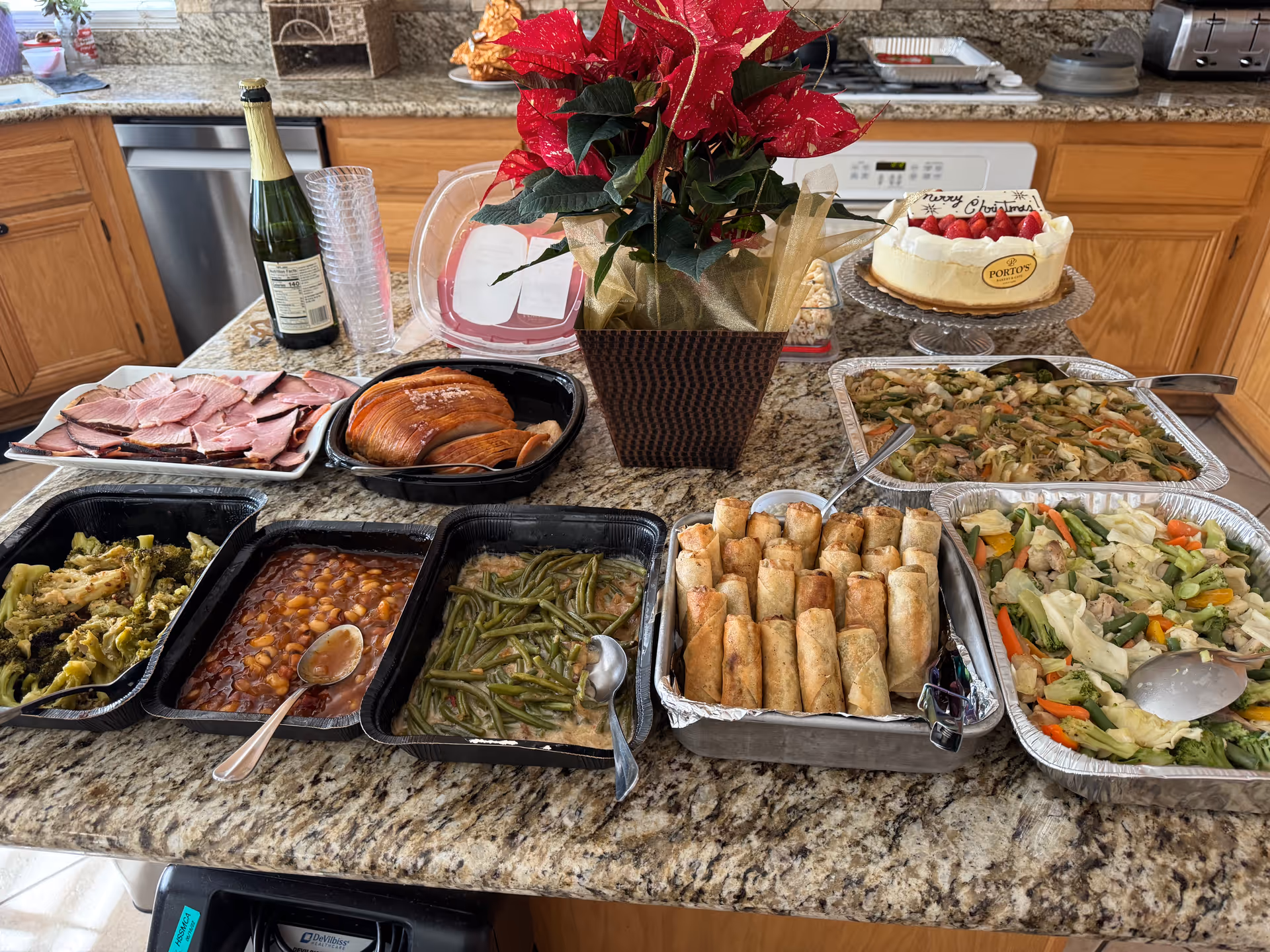 A kitchen countertop with a variety of prepared foods including sliced ham, roasted meat, cooked vegetables, baked beans, green beans, spring rolls, and mixed vegetable dishes. There is a bottle of sparkling beverage with plastic cups, a poinsettia plant in a decorative pot, and a cake with a 'Merry Christmas' message on a glass cake stand. Wooden cabinets and kitchen appliances are visible in the background.