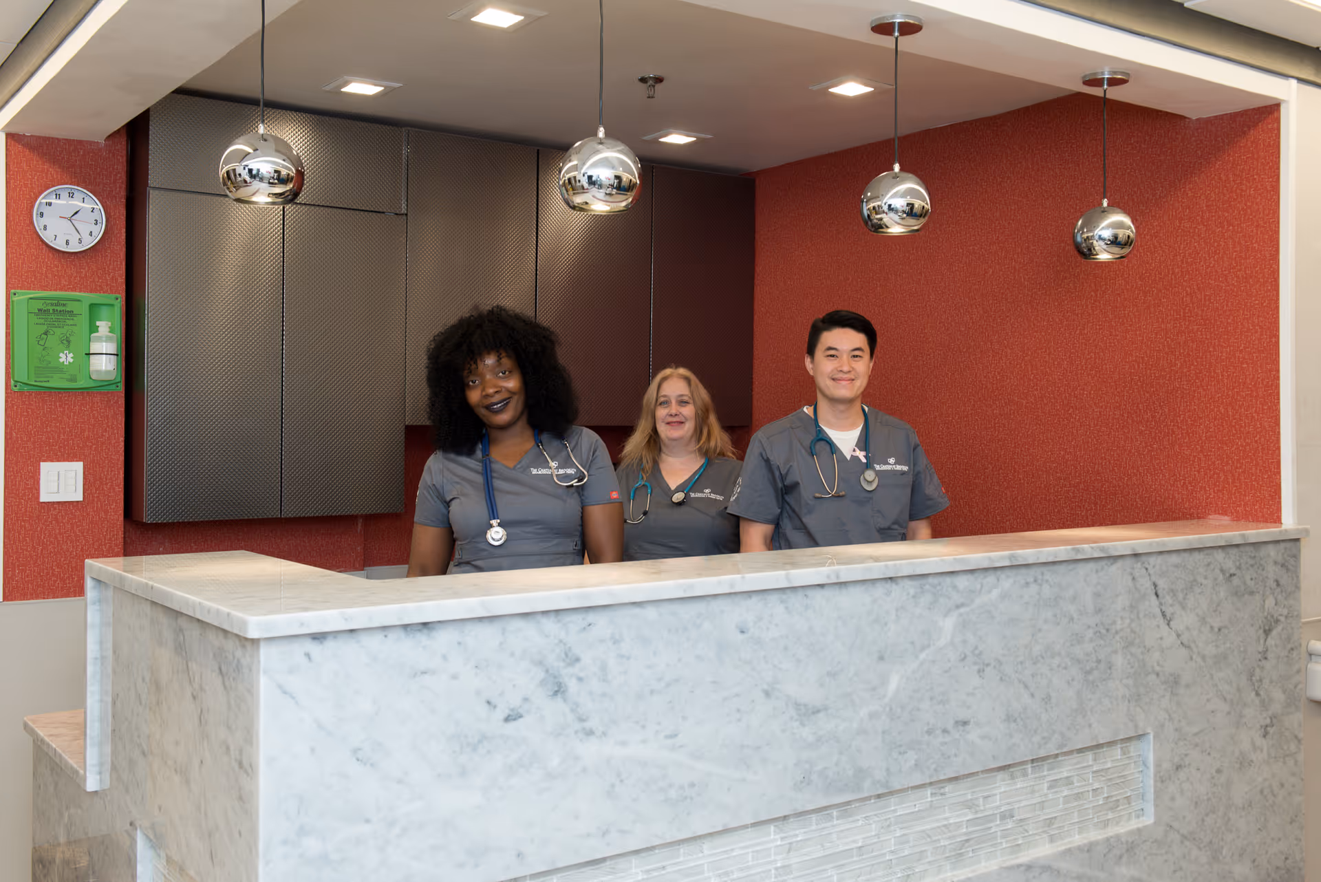 Three healthcare professionals standing behind a marble reception desk in a medical facility. They are wearing gray scrubs and stethoscopes, smiling at the camera. The background features a red textured wall, dark cabinets, a clock, and a green emergency station box.