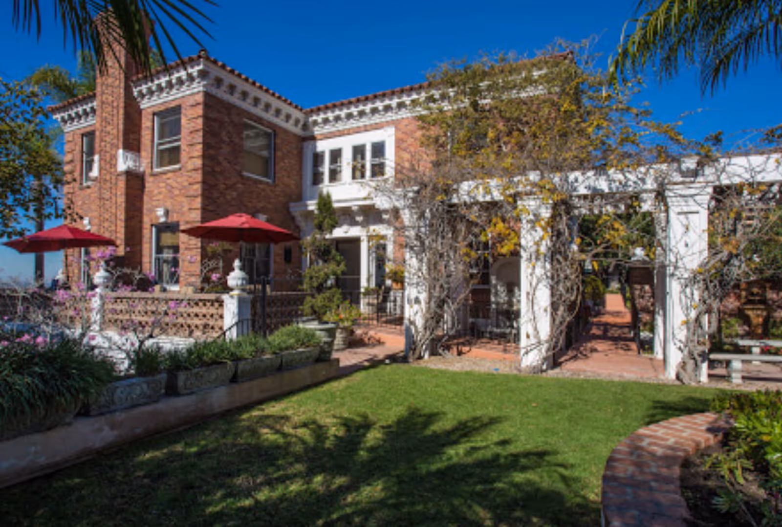 Two-story brick building with a pergola-covered patio, red umbrellas, planters and a grassy front lawn under a clear blue sky.