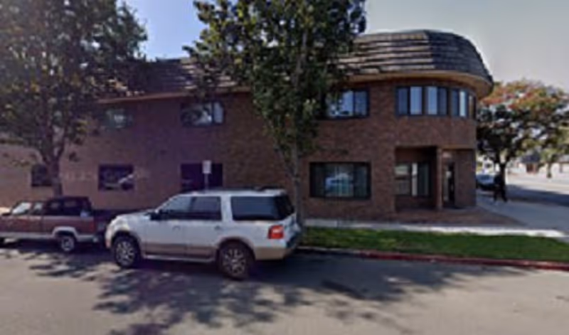Brick building with rounded corner entrance, two parked cars, and trees along a street.