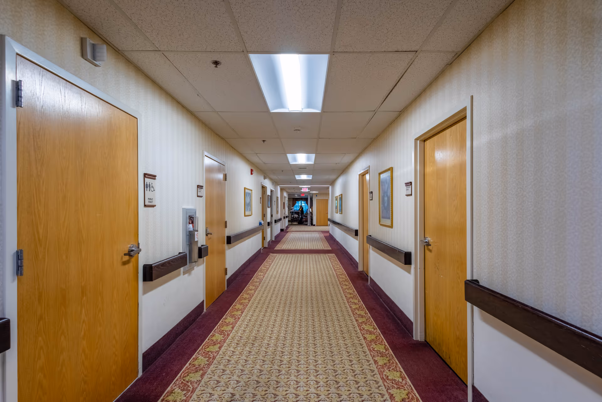 Long carpeted hallway inside a senior living facility with wooden doors on both sides and overhead fluorescent lights.