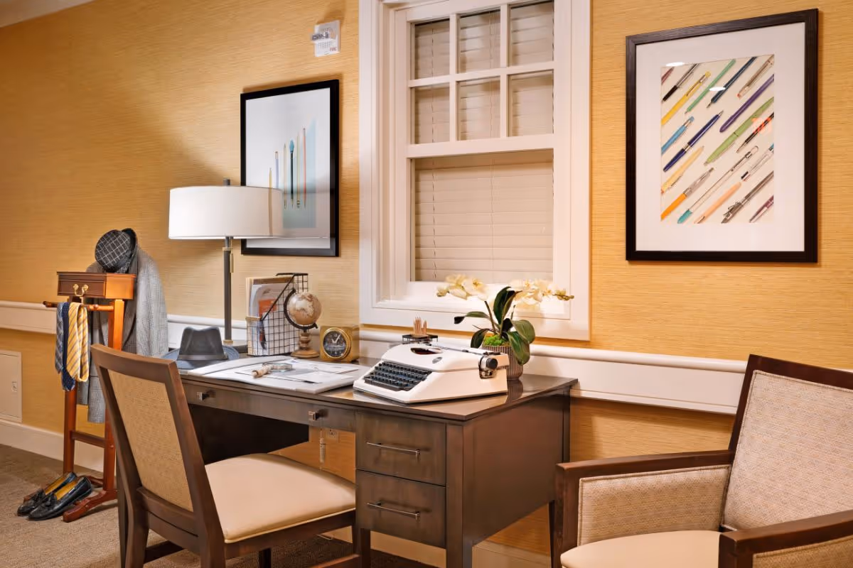 A cozy interior workspace with a wooden desk holding a vintage typewriter, lamp, globe and plant, flanked by chairs and framed artwork.