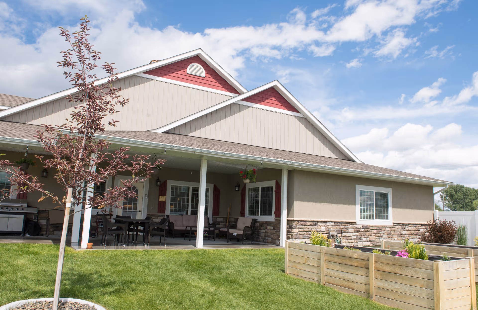 Exterior view of a single-story building with beige and red siding, a covered patio area with outdoor furniture, a small tree in the foreground, and a raised garden bed with plants on a well-maintained lawn under a partly cloudy sky.