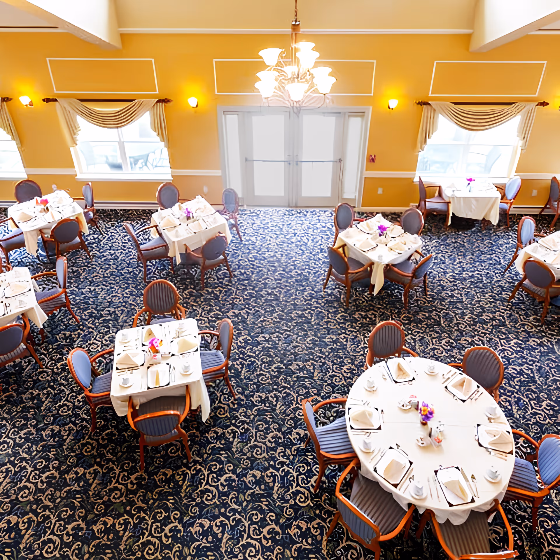 A dining room with multiple round and square tables covered with white tablecloths, set with plates, cups, napkins, and utensils. The room has patterned blue carpet, yellow walls with white trim, large windows with draped curtains, and a chandelier hanging from the ceiling.