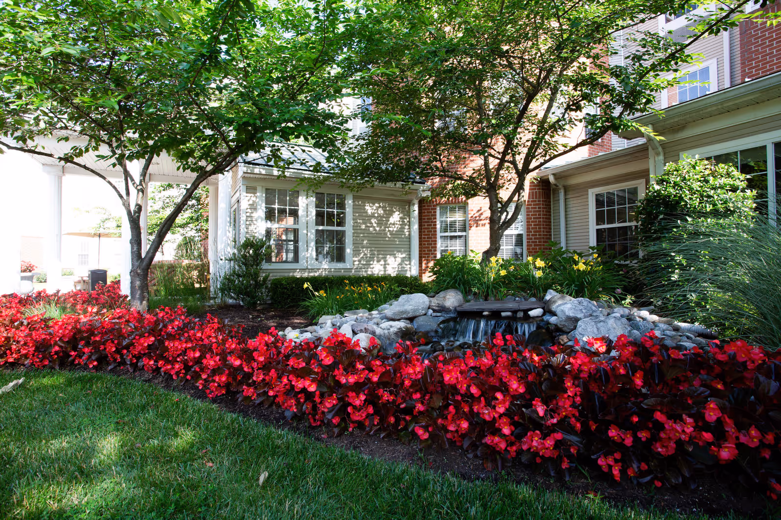 Landscaped front yard with red flowers, a small rock waterfall, trees, and the building's windows.
