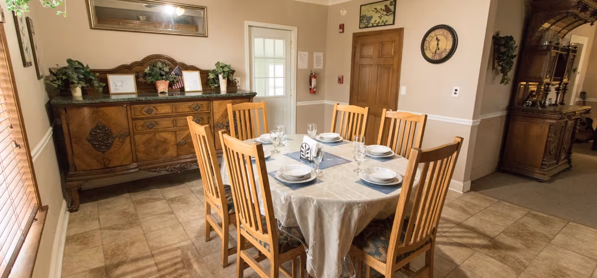 A dining room with a round table covered with a white tablecloth set for six people with plates, glasses, and napkins. The room has wooden chairs around the table, a wooden sideboard with plants and framed items on top, a clock on the wall, and a door with a window. The floor is tiled and the walls are painted beige with white trim.