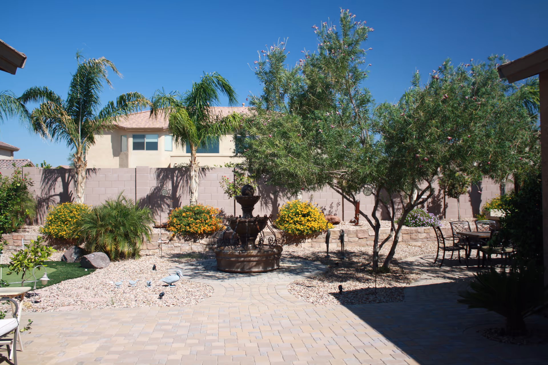 Sunny outdoor courtyard with a central fountain, palm trees, flowering shrubs, and seating.
