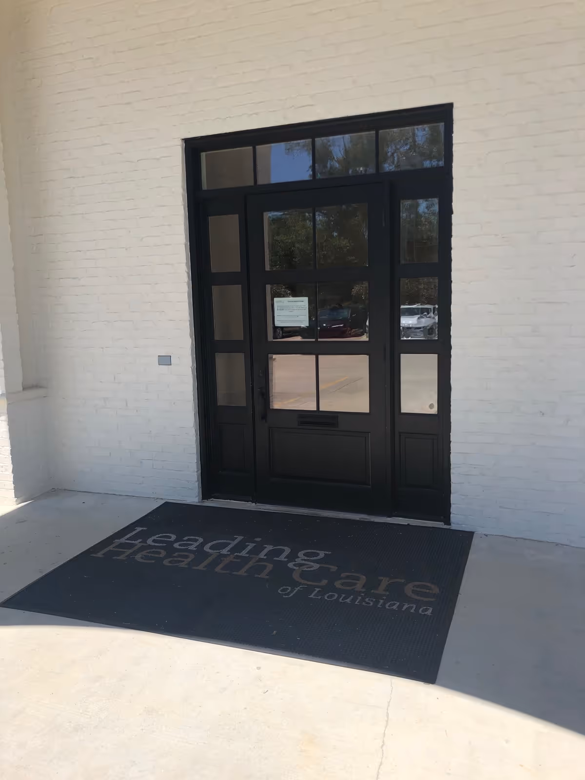 White brick building entrance with a black door and a Leading Health Care of Louisiana mat.