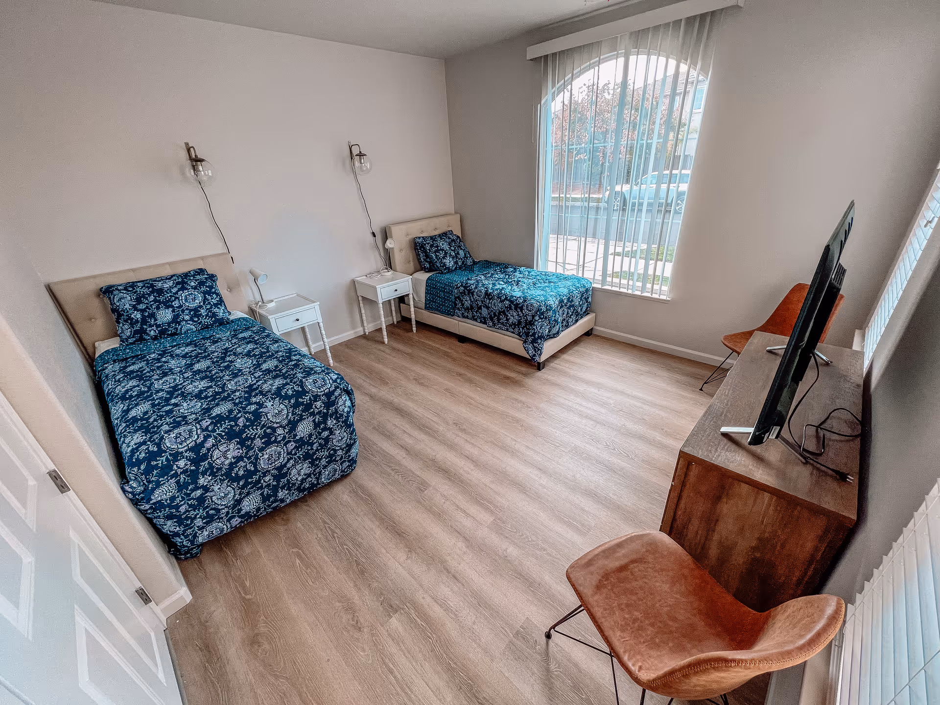 Bright twin bedroom with two single beds in blue patterned bedding, wood-look floors, a TV on a dresser and a large window with vertical blinds.