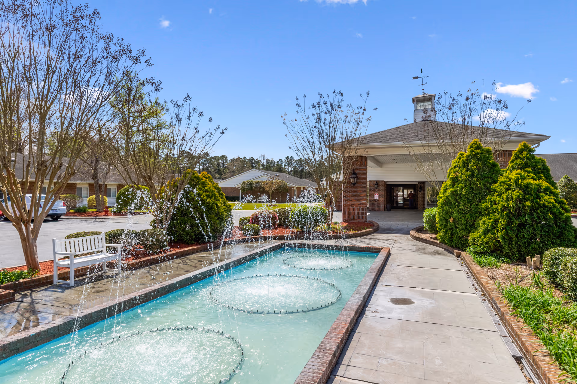 Entrance of a senior living facility with a long rectangular fountain and water jets leading to a covered porte-cochère surrounded by trees and shrubs.