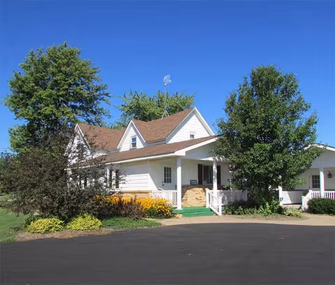 A white house with a brown roof surrounded by green trees and bushes under a clear blue sky. The house has a porch with white railings and a green carpet on the steps leading to the entrance. The driveway in front of the house is paved with asphalt.