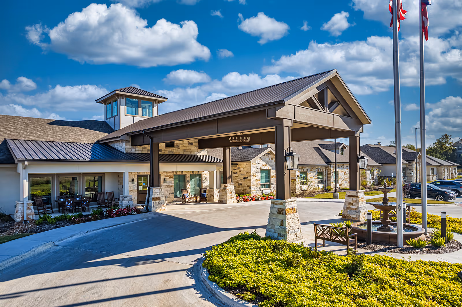 Exterior view of Sage Valley Senior Living facility showing the main entrance with a covered driveway, stone and wood architectural elements, a landscaped roundabout with a fountain, benches, and flagpoles under a blue sky with scattered clouds.