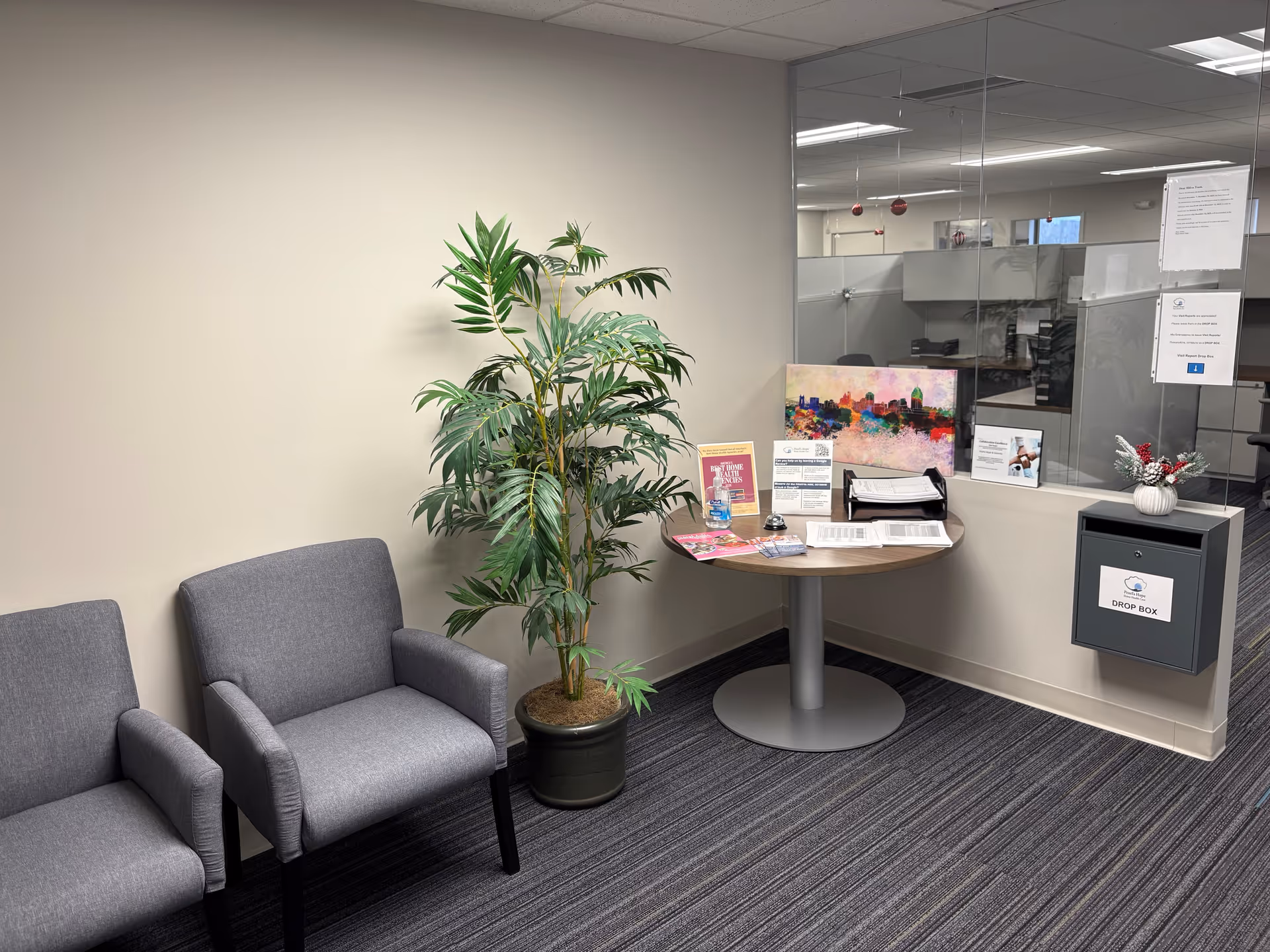 Visitor lobby with two gray chairs, round table, potted plant, glass partition, and drop box.