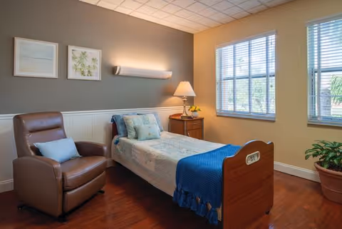 Senior living bedroom with bed, armchair, nightstand, lamp, and large windows.